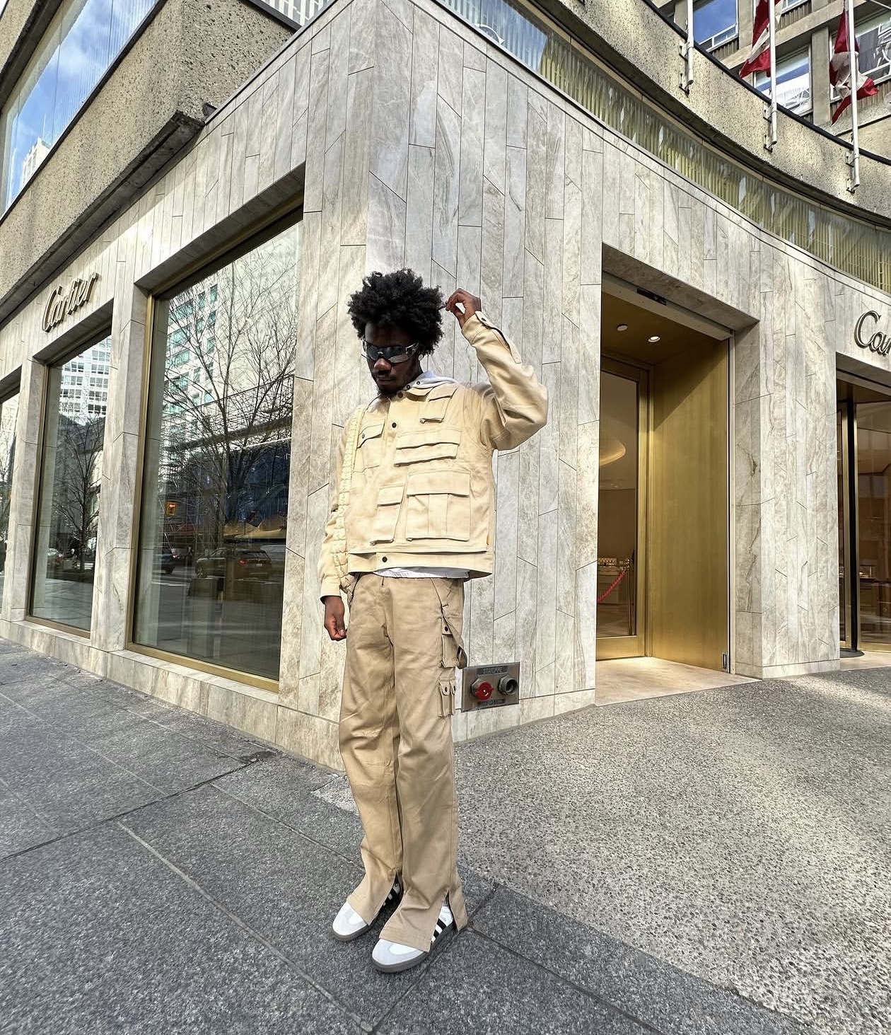 Editorial portrait — beige utility jacket, cargo trousers and sunglasses outside a luxury boutique